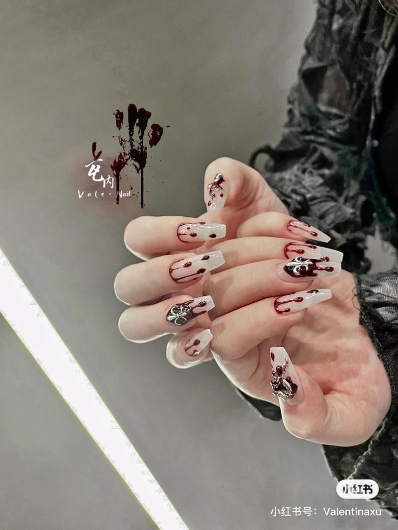 mau-nail-halloween-dep__24__a25d3aecb63b45dca602b4d9f3328cc5 Bàn tay với móng tay dài, trang trí họa tiết màu đỏ và đen trên nền màu trắng, phong cách nghệ thuật.