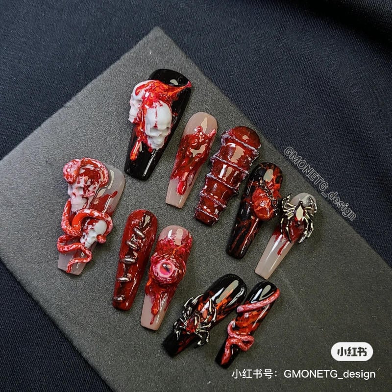 mau-nail-halloween-dep__2__5dcd75462a6844a998457069b7b043cd Bộ móng nghệ thuật với họa tiết nổi hình đầu lâu và nhện, màu đỏ và đen, trên nền vải tối màu.