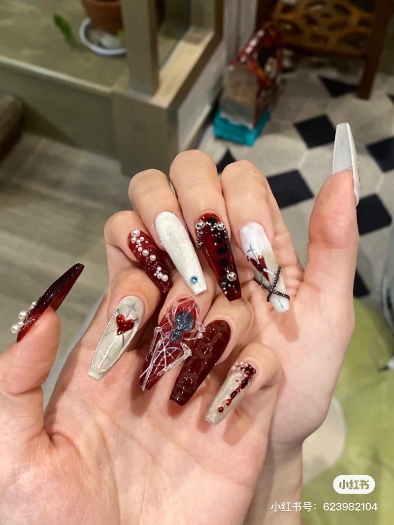 mau-nail-halloween-dep__34__b55b7b39e71d4be5952090dae23853ba Bàn tay với móng tay dài, được trang trí cầu kỳ với các họa tiết và màu sắc đa dạng như đỏ, trắng và bạc.