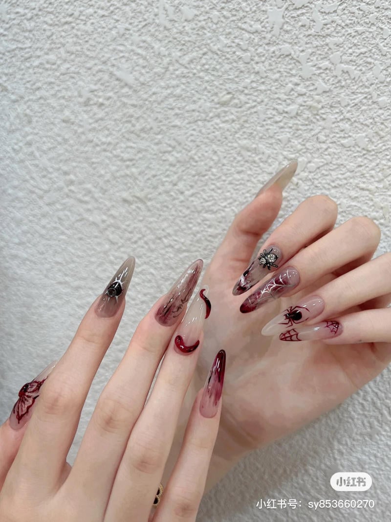 mau-nail-halloween-dep__5__3e48f6504cfc448d83d6065759088d2d Bàn tay với móng tay dài, được trang trí họa tiết chủ đề Halloween, bao gồm nhện và mạng nhện, trên nền tường trắng.