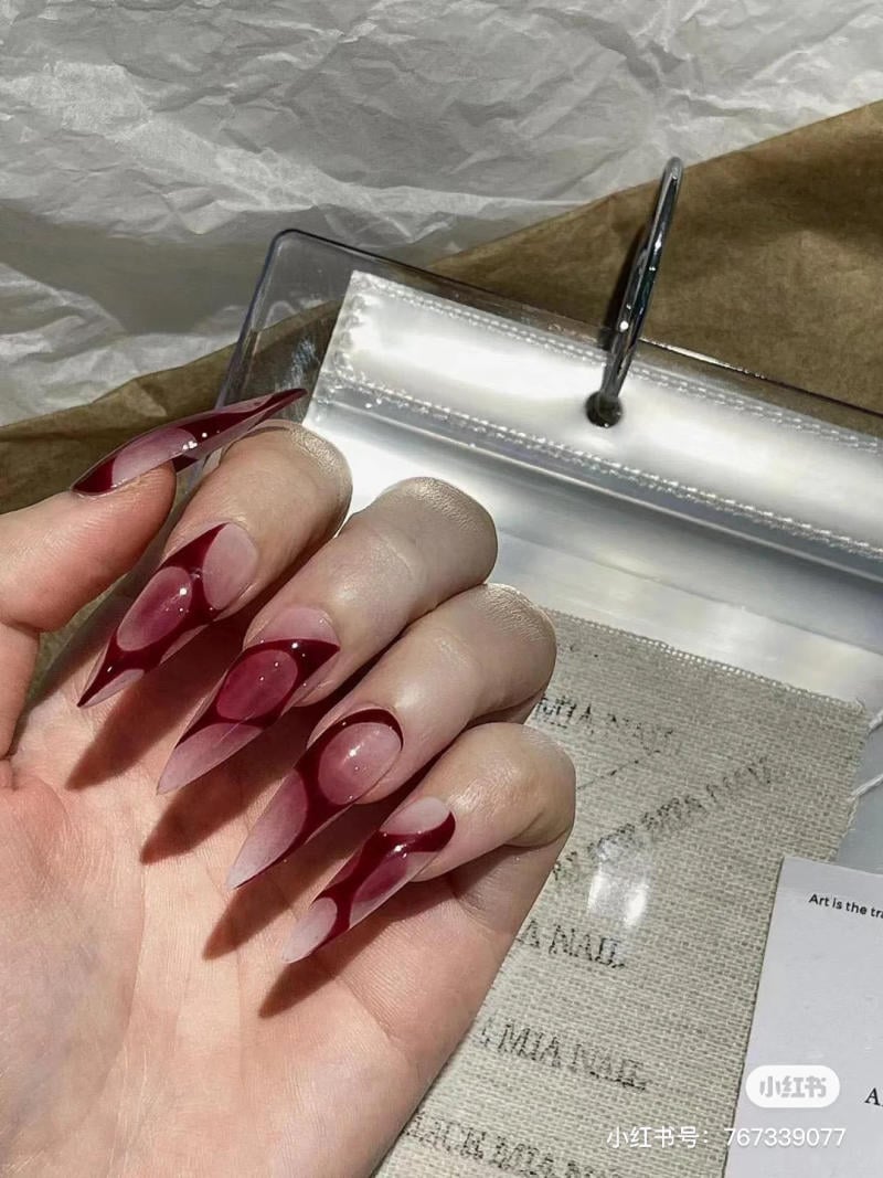mau-nail-halloween-dep__69__ca0209ff3d03440f941e80d8c5239bc3 Bàn tay với móng tay dài, sơn họa tiết đỏ trên nền màu nhạt, đặt trên nền giấy có chữ và bìa nhựa trong suốt.