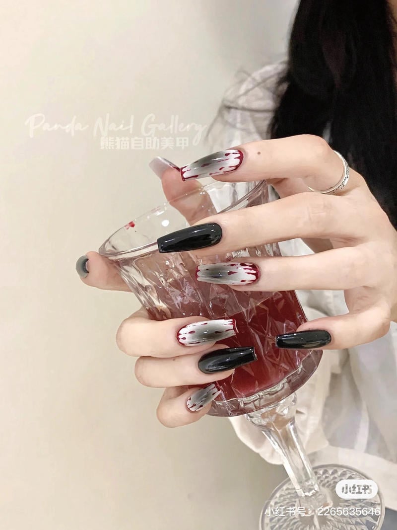 mau-nail-halloween-dep__6__e72e00c4971f4739b3821acfe58e8fb9 Bàn tay với móng tay sơn đen và đỏ, cầm ly thủy tinh đựng chất lỏng màu đỏ.