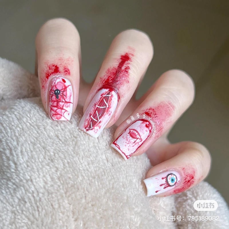 mau-nail-halloween-dep__8__ed717a38c14a44548bc7065fa6ca06c8 Móng tay được sơn nghệ thuật với họa tiết mạng nhện, mắt và vết đỏ trên nền trắng, tạo phong cách độc đáo.