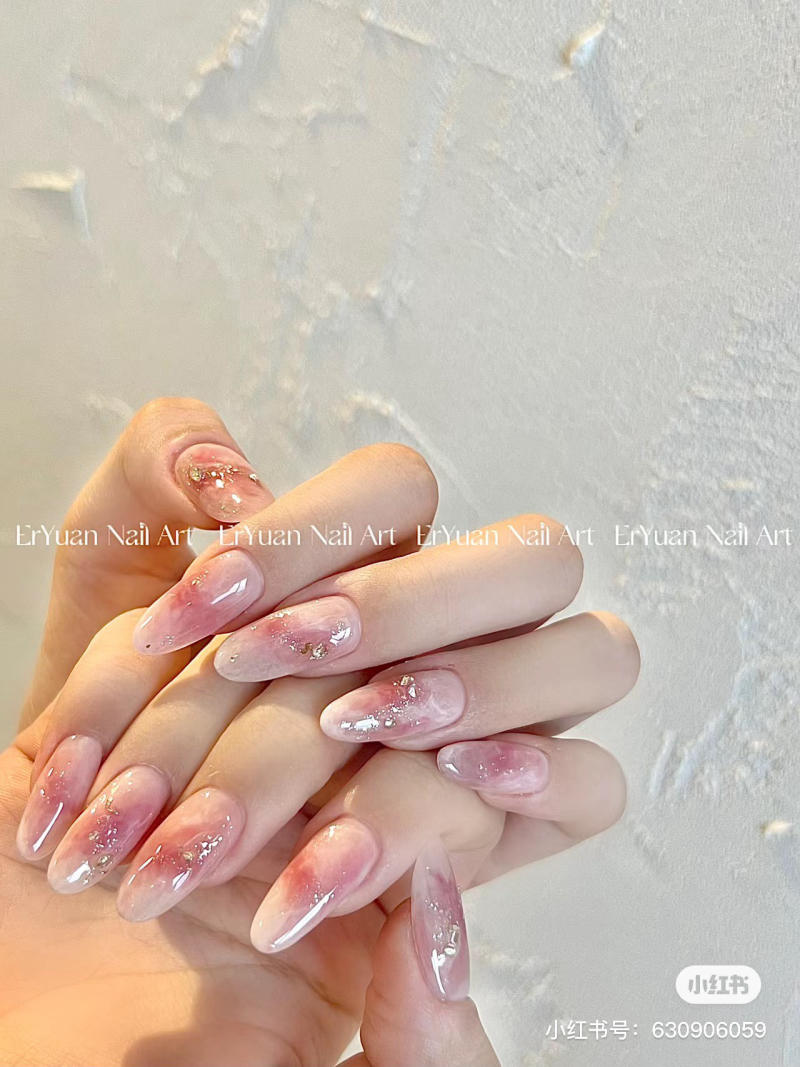 Bàn tay với móng tay được sơn màu hồng nhạt, trang trí hoa văn ánh kim, nền tường trắng.