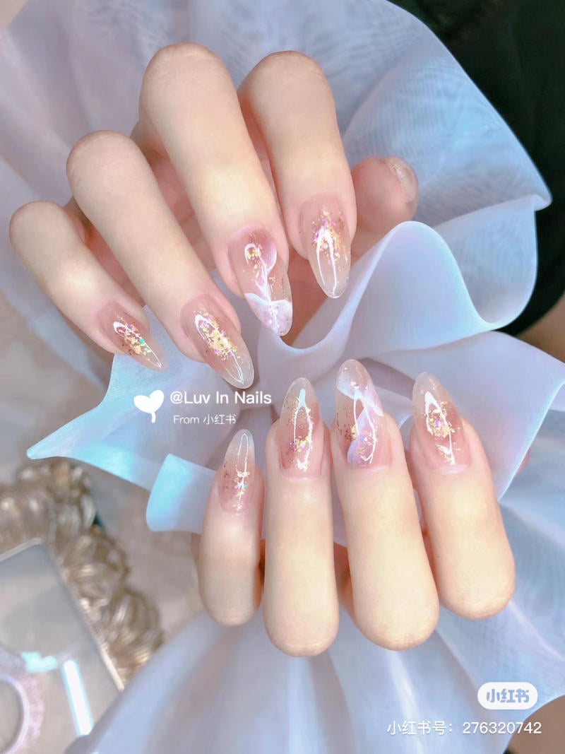 Bàn tay với móng tay được trang trí họa tiết lấp lánh và ánh kim, nền vải trắng nhẹ nhàng phía sau.