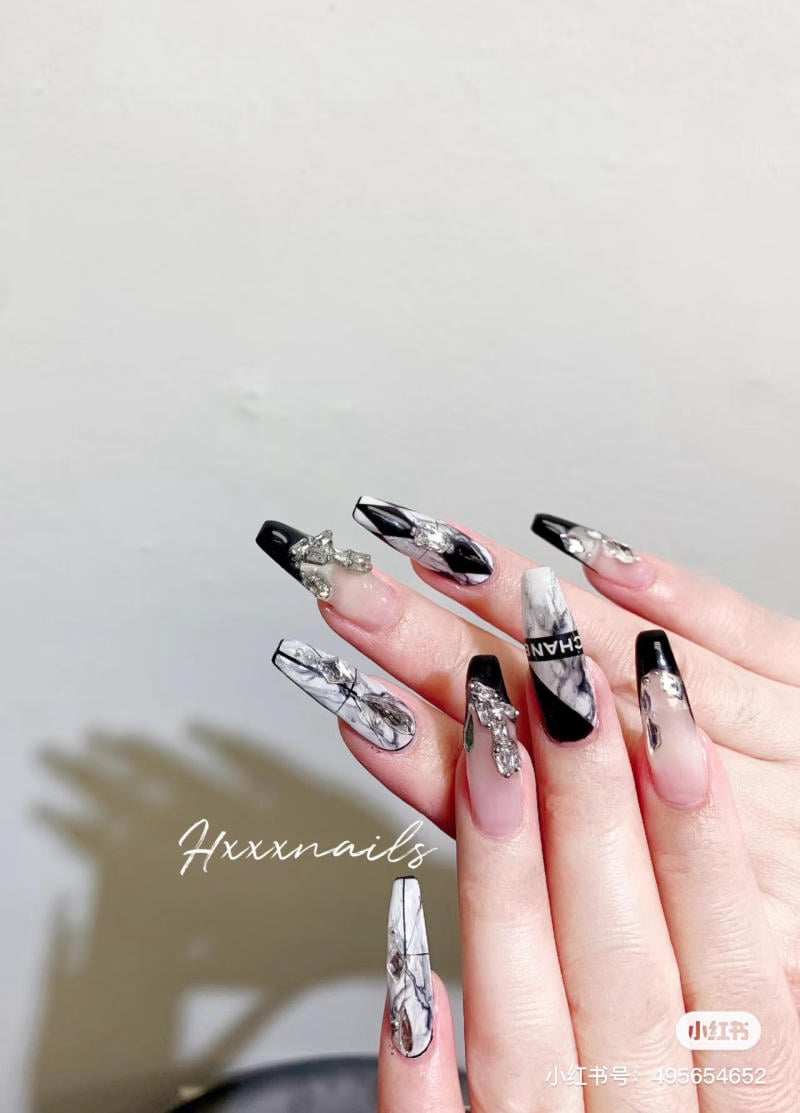 Bàn tay với móng tay dài, trang trí hoa văn đen trắng và đính phụ kiện kim loại sáng bóng.