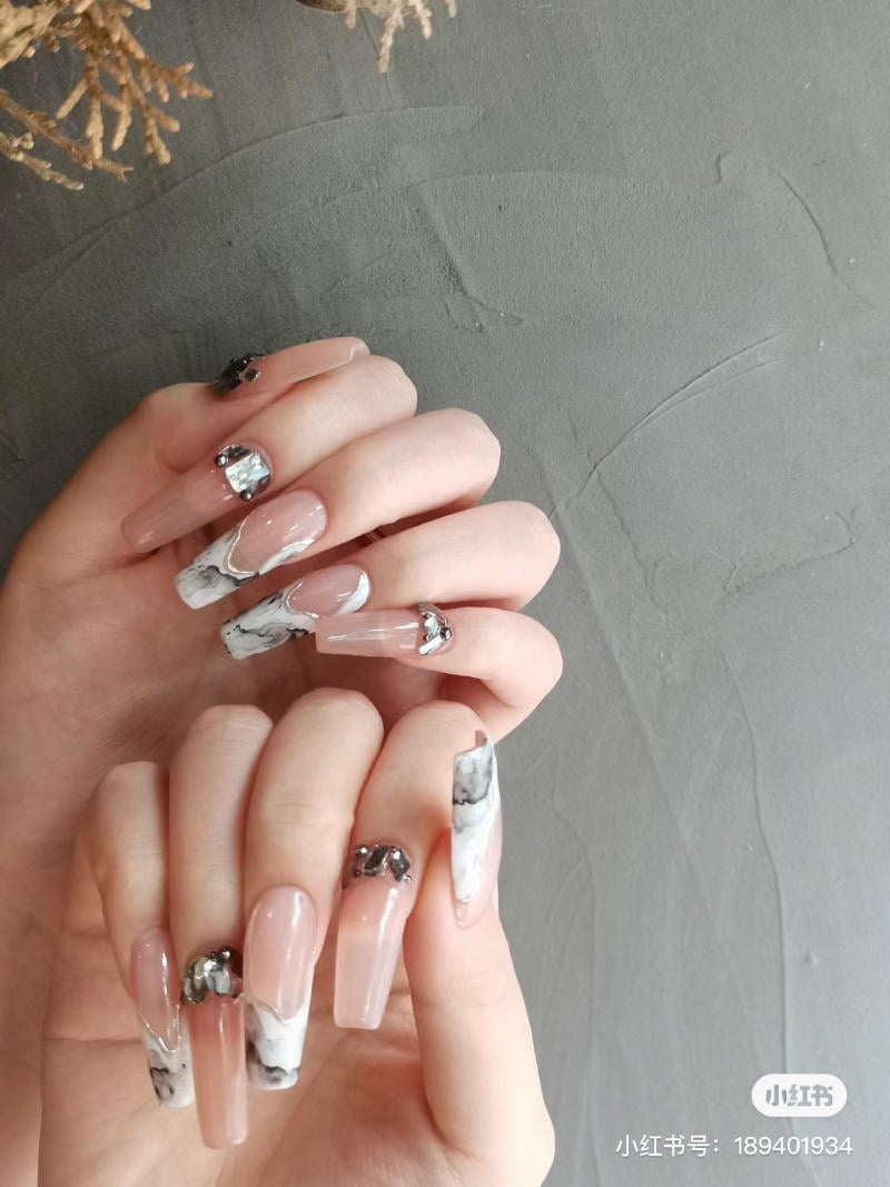 Bàn tay với móng dài được trang trí bằng họa tiết đá và hoa văn đá cẩm thạch trên nền xám nhạt.