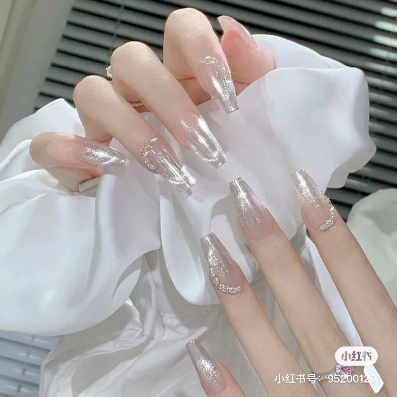 Bàn tay với móng tay dài được sơn màu bạc lấp lánh, trên nền vải trắng.