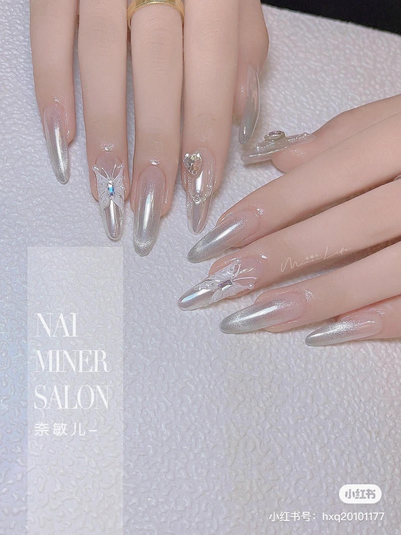 Bàn tay với móng tay nhọn, phủ sơn màu bạc, trang trí họa tiết bướm và đá lấp lánh.