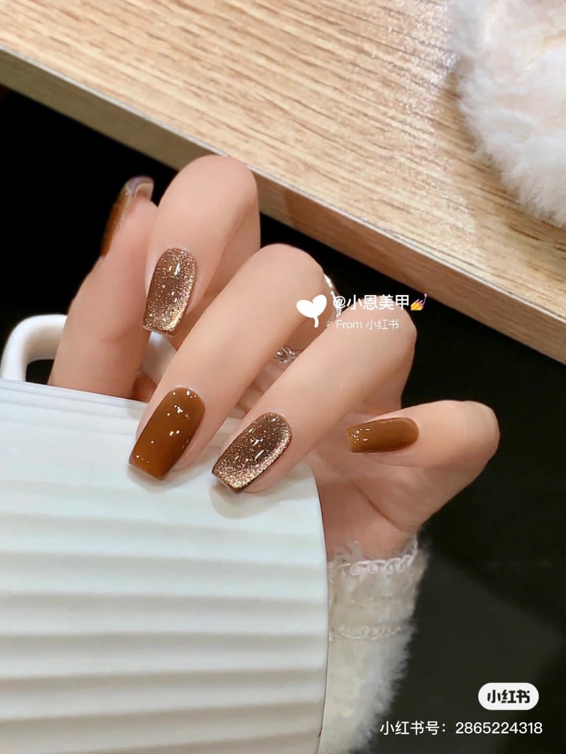 Bàn tay với móng màu nâu và lấp lánh, cầm một chiếc cốc trắng, nền là mặt bàn gỗ.
