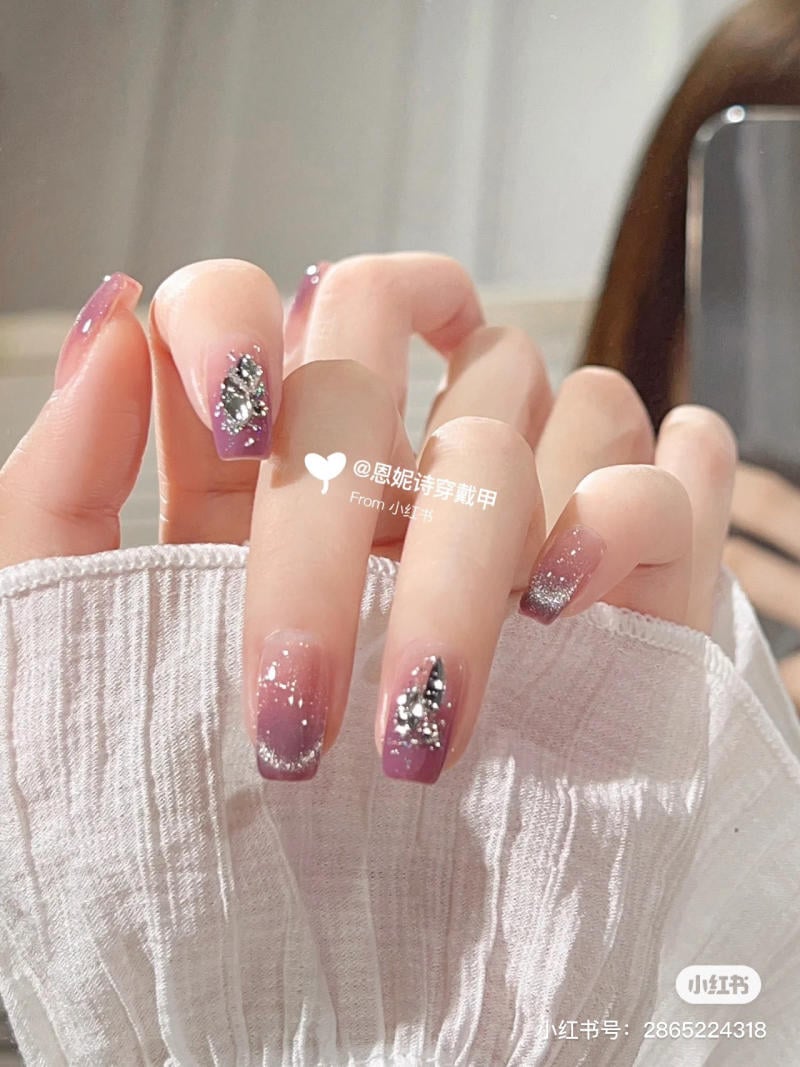 Bàn tay với móng tay được trang trí lấp lánh, sơn màu tím nhạt, có họa tiết kim cương và đường kim tuyến.