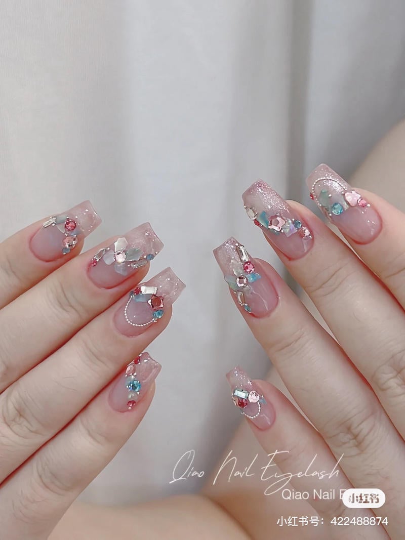 Bàn tay với móng tay dài, trang trí đá và hoa văn tinh tế trên nền màu hồng nhạt.