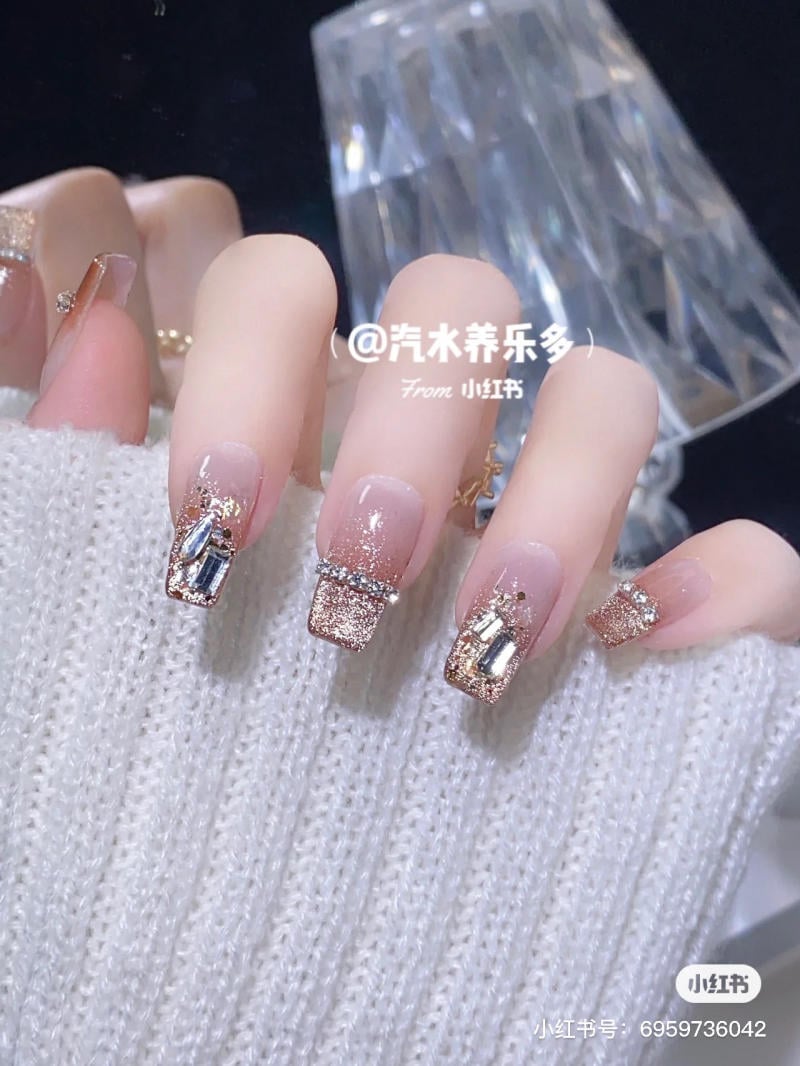 Bàn tay với móng tay sơn màu hồng nhạt, trang trí kim tuyến và đá lấp lánh, trên nền áo len trắng.