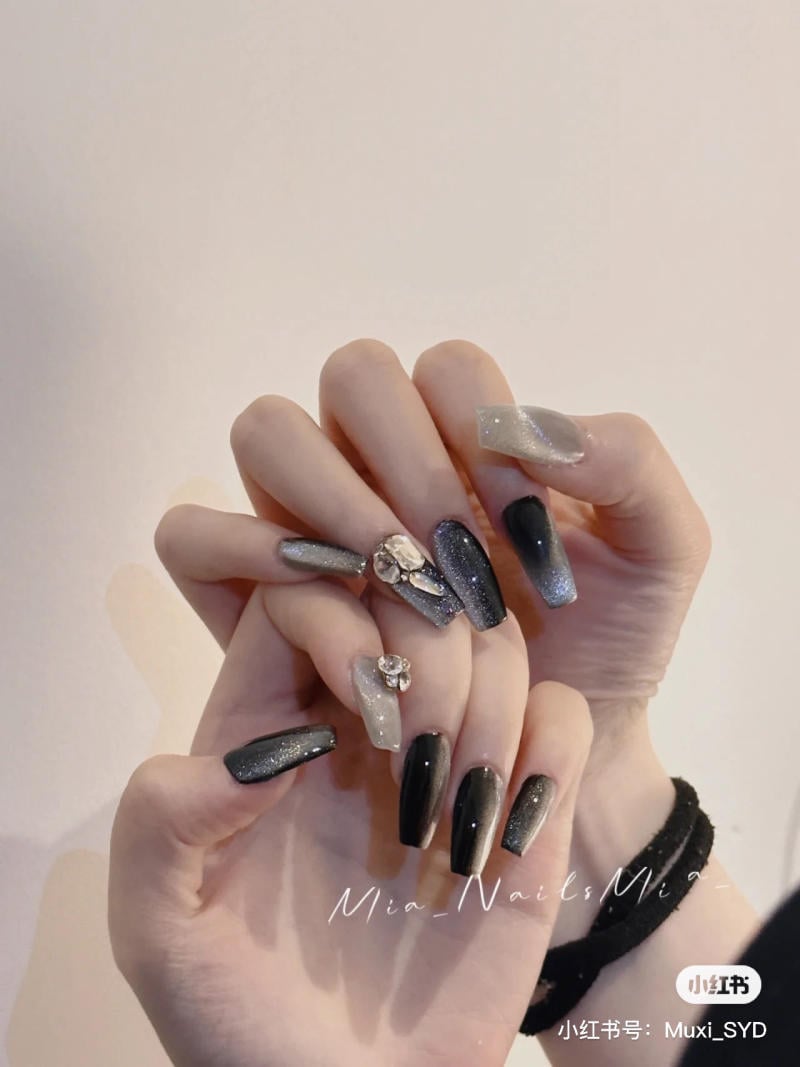 Bàn tay với móng tay dài, sơn màu đen và xám ánh kim, trang trí đá lấp lánh trên nền trắng.