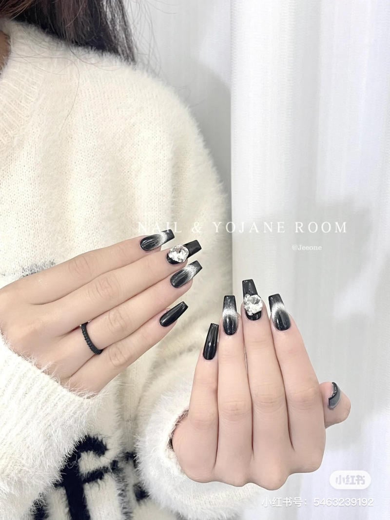 Bộ móng tay sơn màu đen bóng, trang trí đá và kim tuyến, người mẫu mặc áo len trắng.