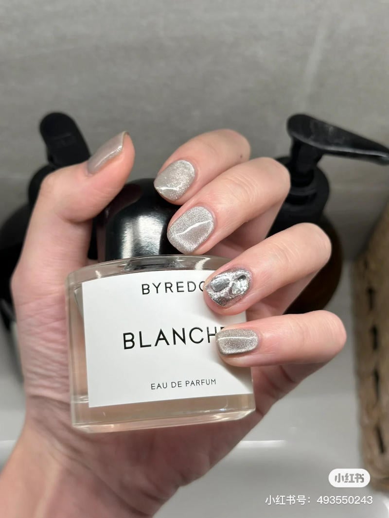 Bàn tay cầm chai nước hoa, móng tay được sơn màu bạc lấp lánh với họa tiết trang trí.