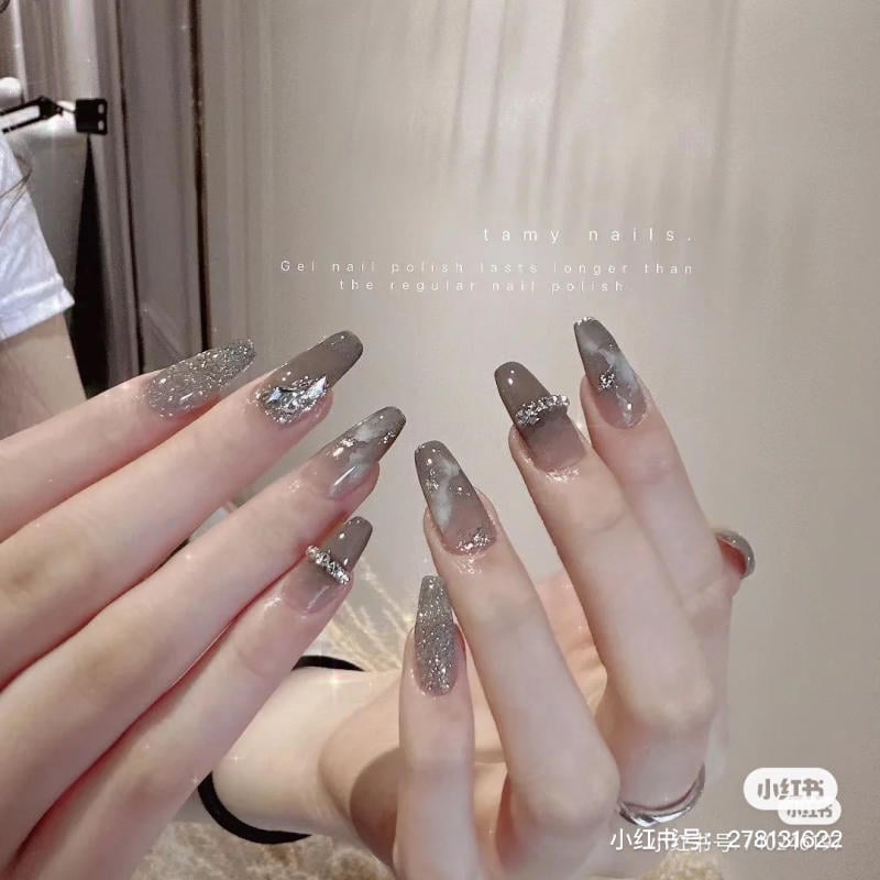 Bàn tay với móng tay được sơn gel màu xám, trang trí lấp lánh và đính đá, đang được chụp cận cảnh.