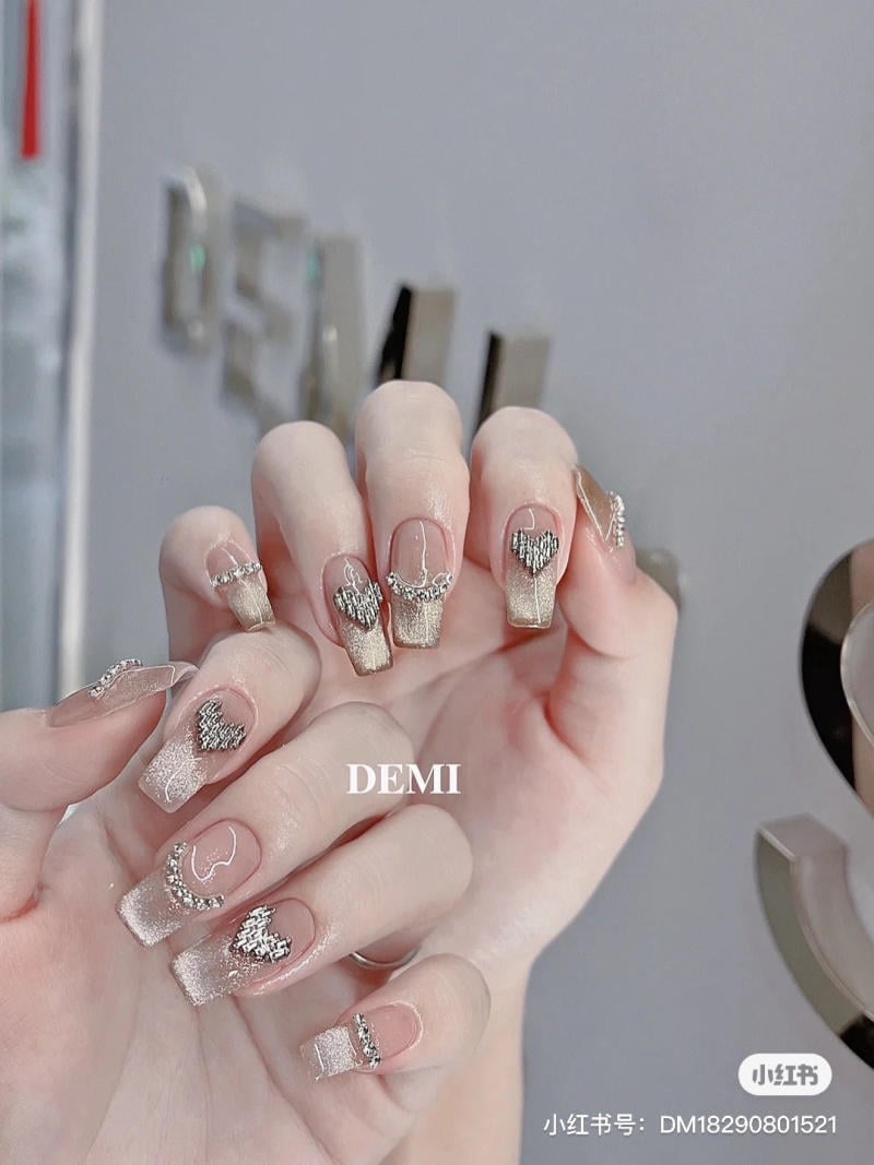 Bàn tay với móng tay dài được trang trí bằng họa tiết trái tim và đính đá lấp lánh trên nền màu nhạt.