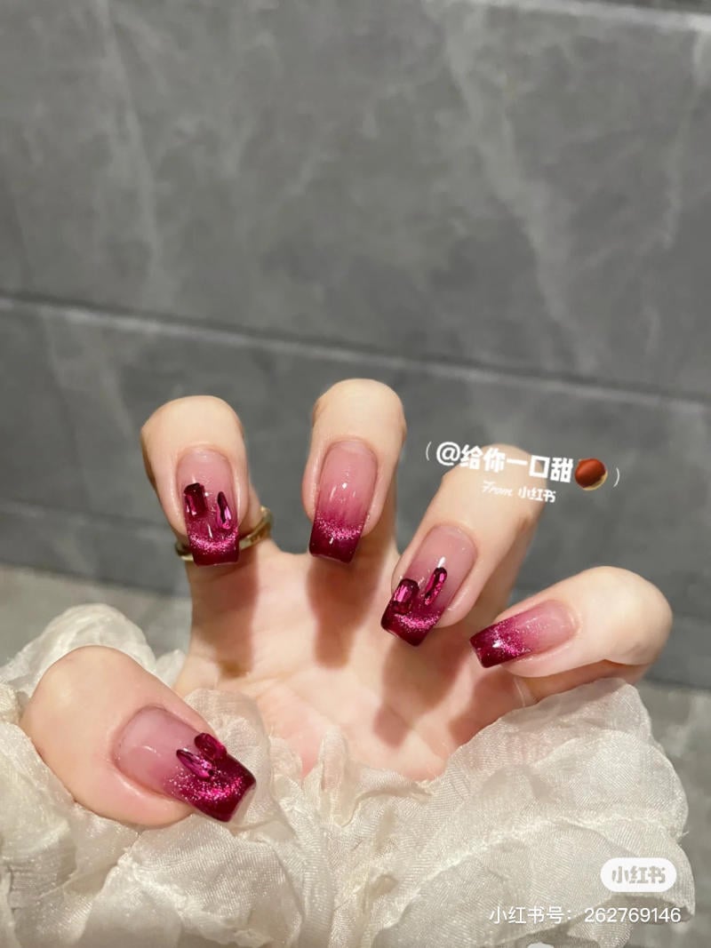 Bàn tay với móng tay dài, sơn màu đỏ đậm, nhấn nhũ lấp lánh trên nền gạch xám.