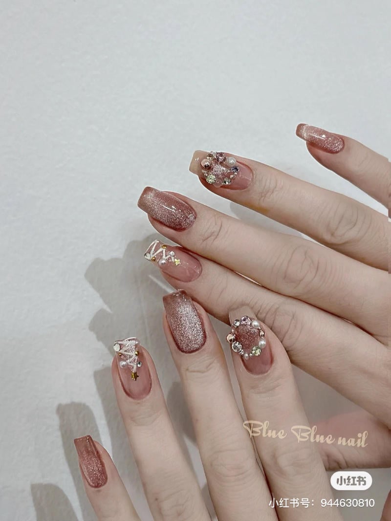 Bàn tay với móng tay dài, sơn màu hồng nhũ, trang trí đá và hoa văn sáng tạo.