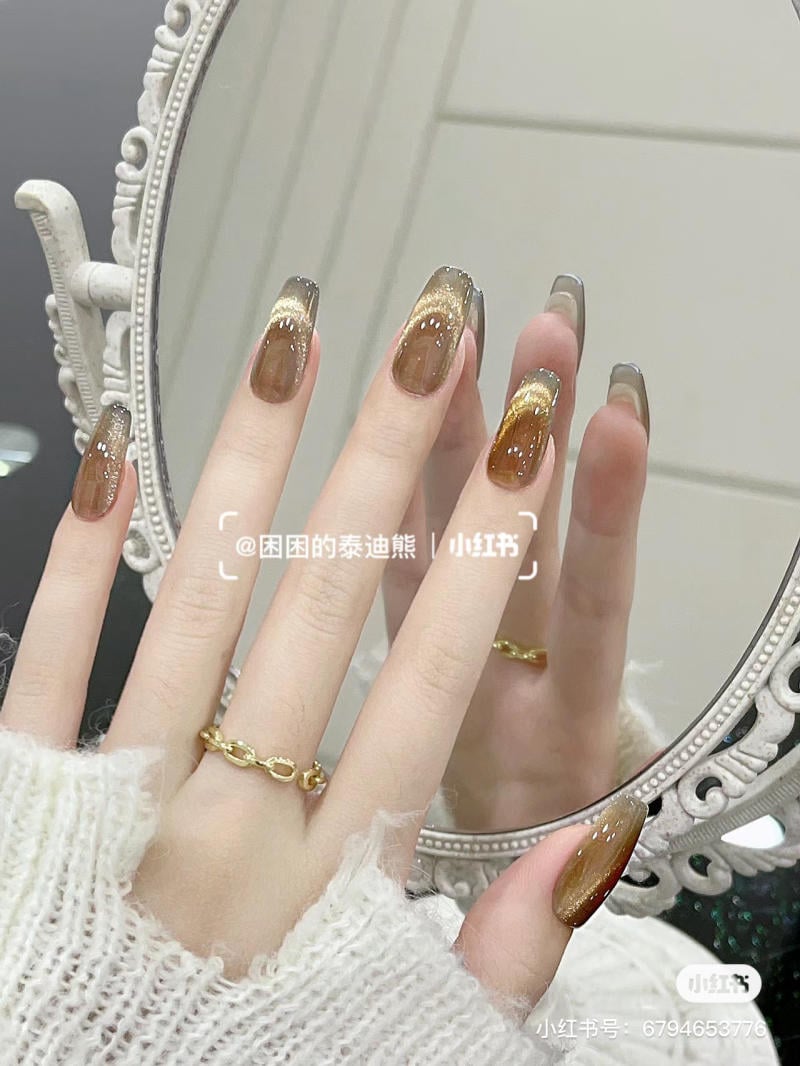Một bàn tay đeo nhẫn vàng, móng tay được sơn màu nâu sẫm và bóng, đặt trước gương trang trí.