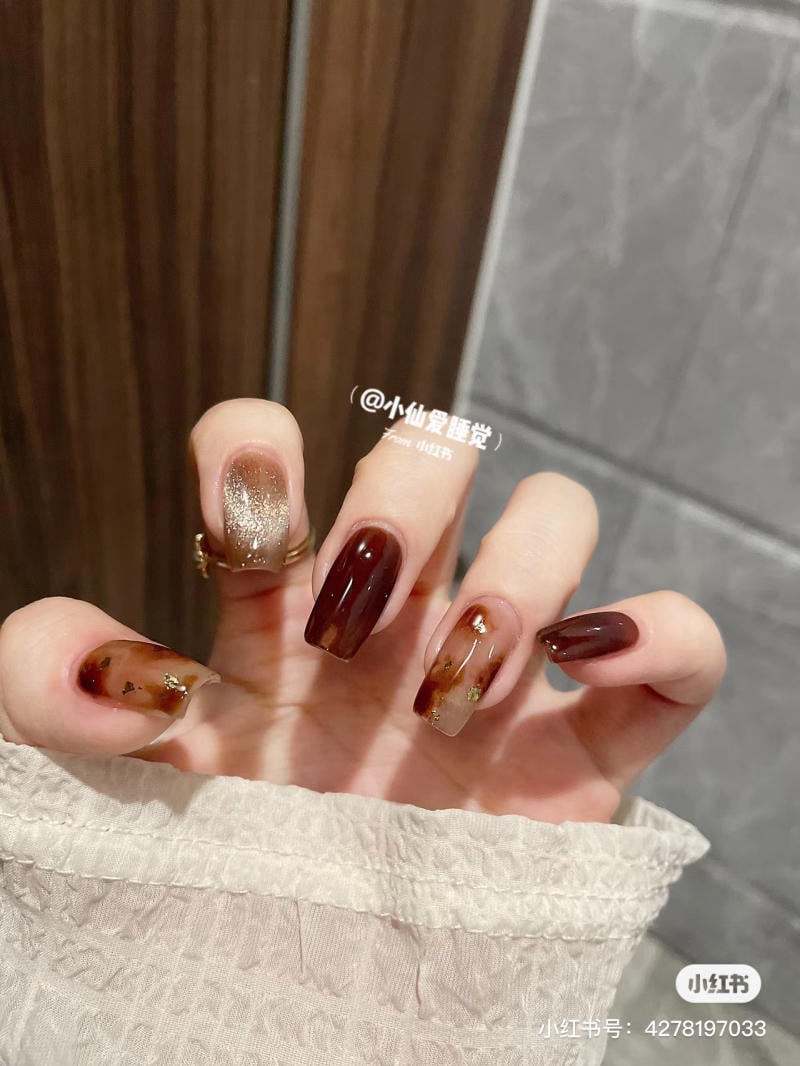 Bàn tay với móng tay sơn màu nâu và vàng lấp lánh, nền gạch và gỗ.