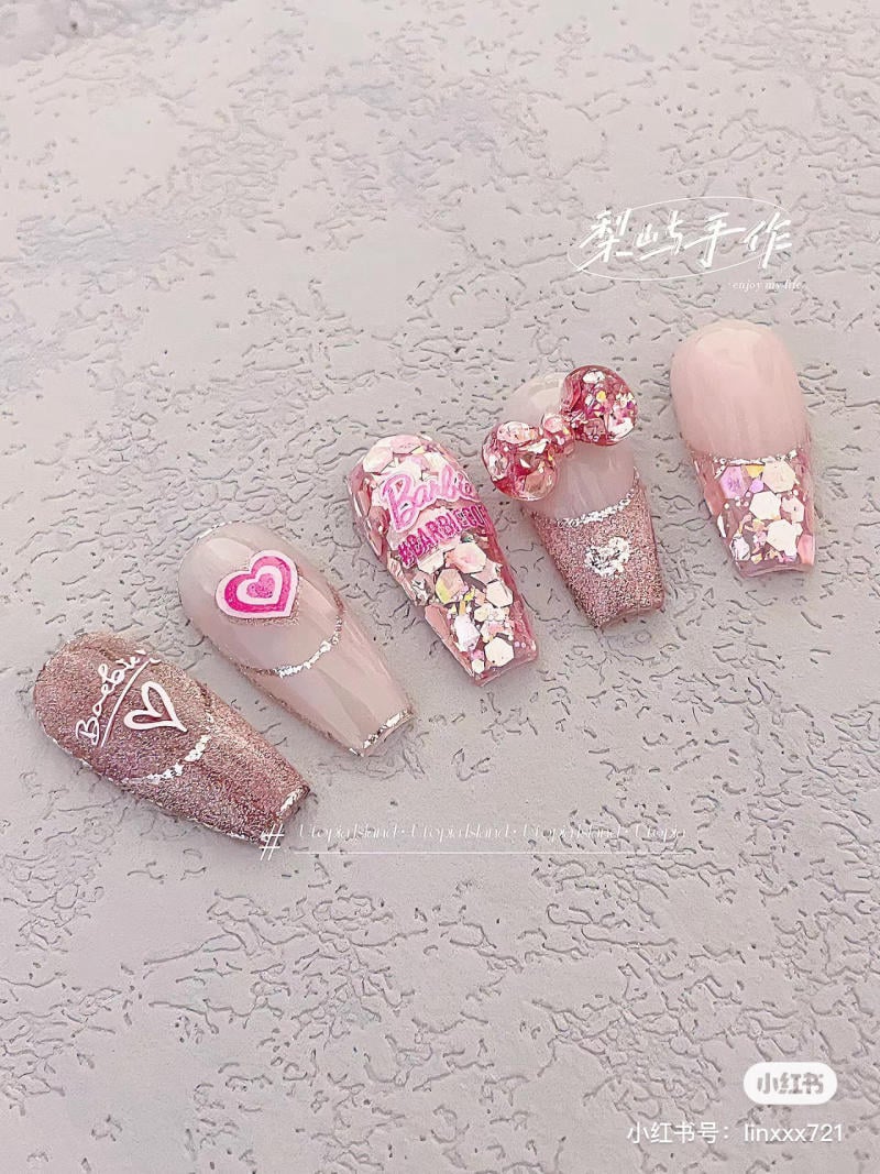 Bộ móng tay giả màu hồng nhạt, có hoa văn trái tim, chữ và đính đá lấp lánh trên nền xi măng xám.