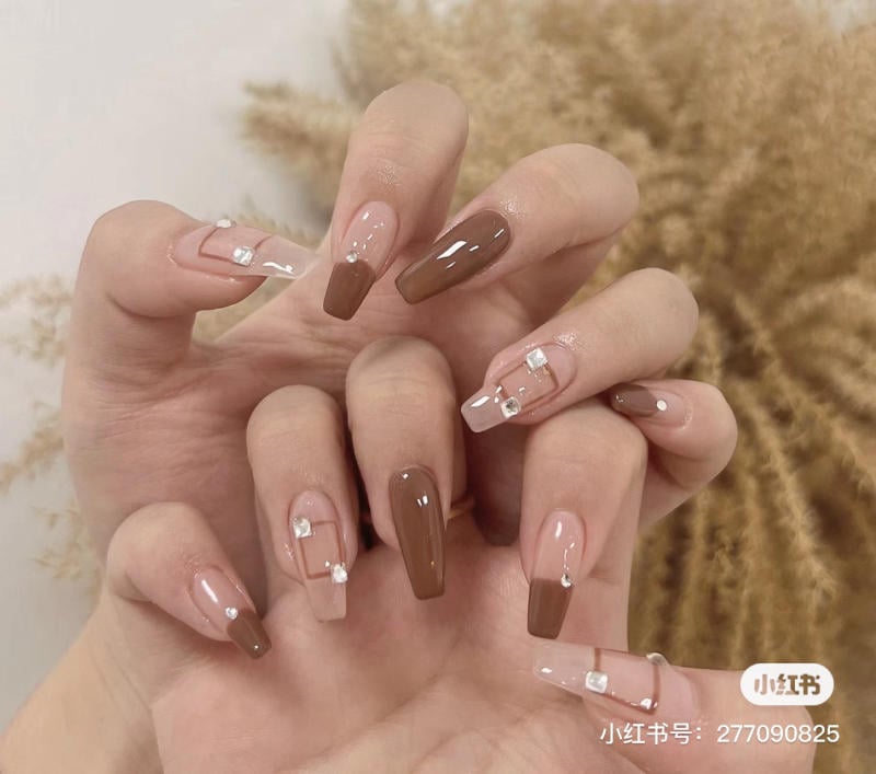 Bàn tay với móng tay sơn màu nâu và hồng nhạt, trang trí đá nhỏ, nền mờ với cây khô phía sau.