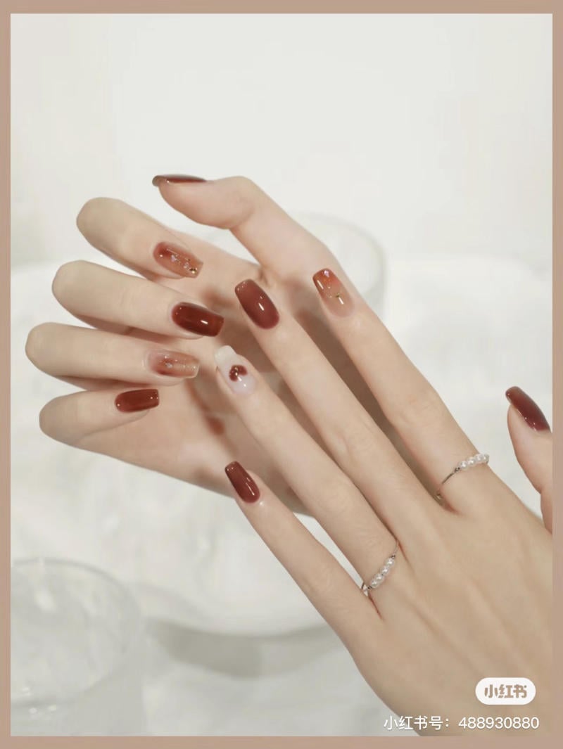 Bàn tay với móng tay sơn màu đỏ và nâu, đeo nhẫn bạc, nền trắng nhẹ nhàng.