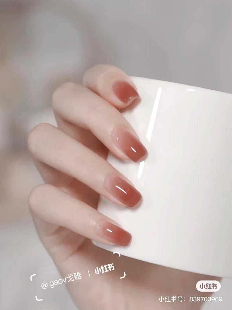 Bàn tay cầm cốc trắng, móng tay sơn màu nâu bóng, nền mờ.