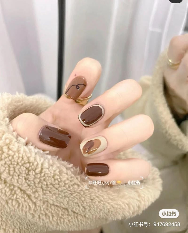 Bàn tay với móng tay sơn màu nâu và be, có họa tiết trang trí, đang cầm một chiếc áo lông màu be.