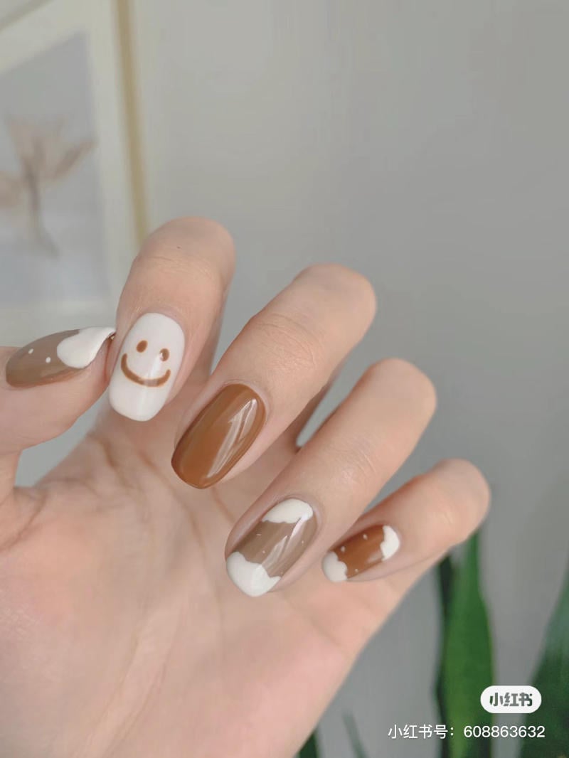 Bàn tay với móng tay được sơn màu nâu và trắng, có họa tiết mặt cười và đám mây.