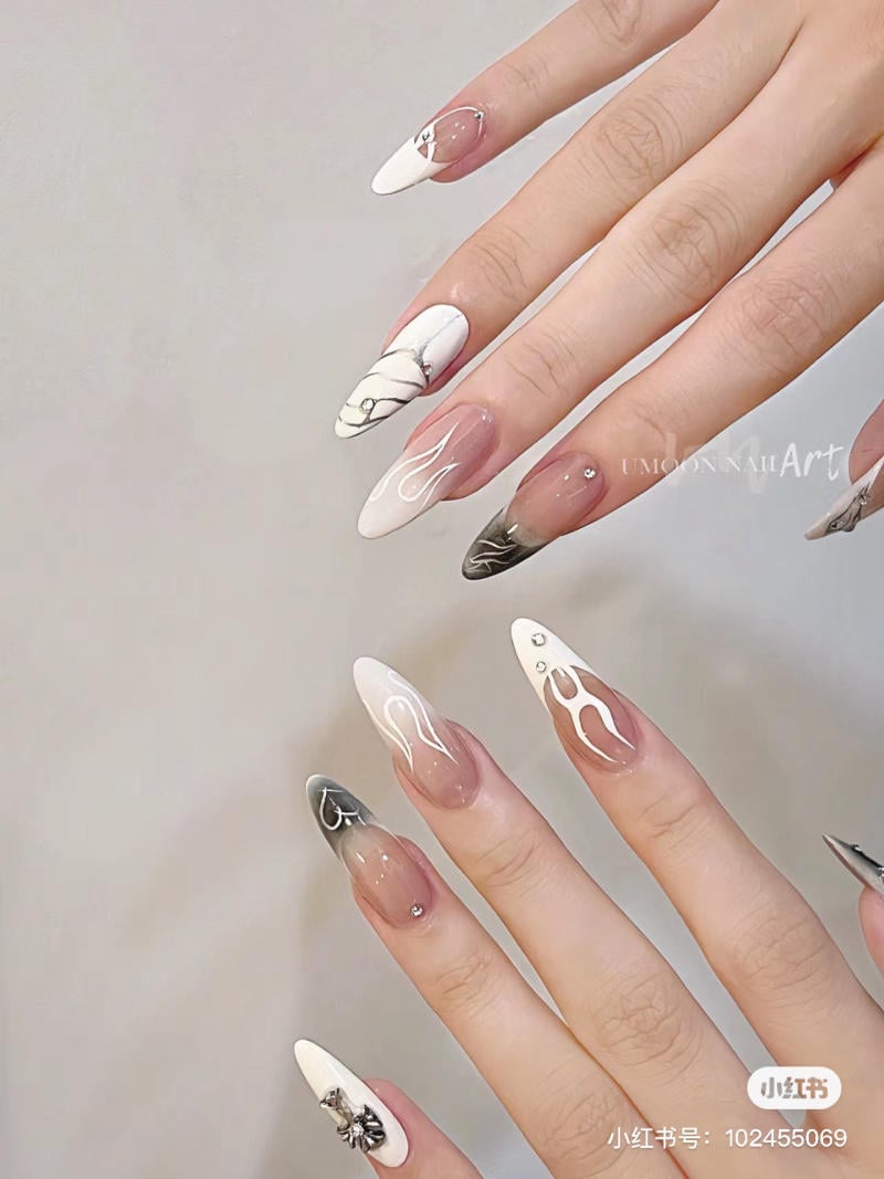 mau-nail-mong-nhon-dai__10__cb5f0858e60246b1aea5330413915b16 Bàn tay với móng tay được trang trí nghệ thuật, có họa tiết trắng, đen và kim loại trên nền màu nude.