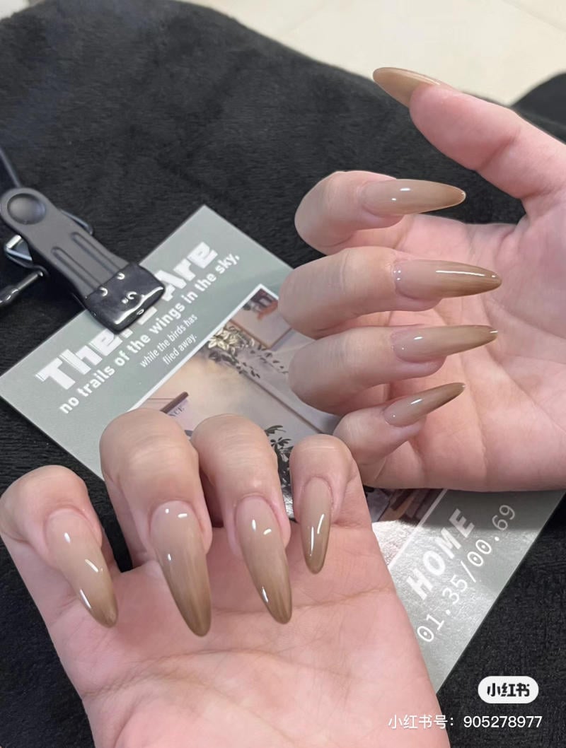 mau-nail-mong-nhon-dai__11__92206f6d4bfe41188e6c7eeaad9f90cc Bàn tay có móng tay dài, sơn màu nâu bóng, cầm cuốn tạp chí trên nền vải màu đen.