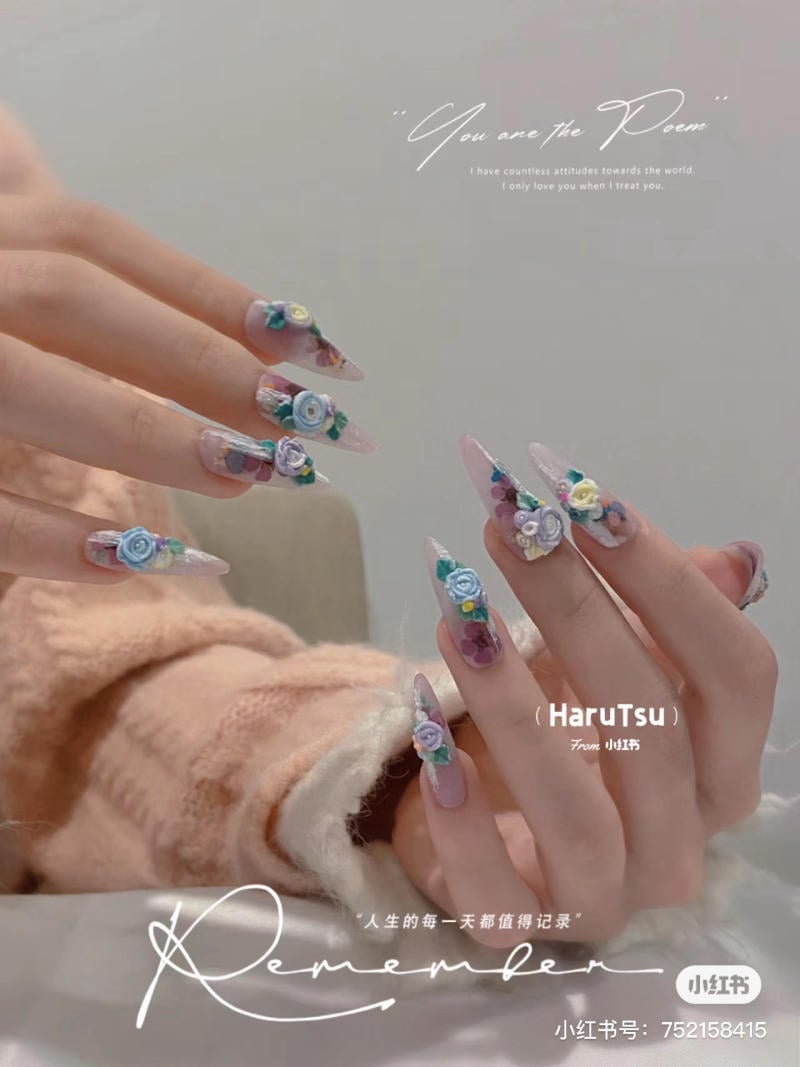 mau-nail-mong-nhon-dai__13__86c0987b7796423bbf72e0b03eba6818 Bàn tay với móng tay dài được trang trí hoa nổi, nền áo len màu hồng nhạt. Văn bản trang trí trên hình.