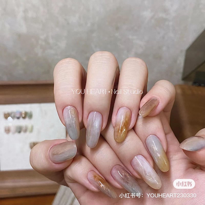 mau-nail-mong-nhon-dai__3__69b48217f7a64ef8a2b6275f28989476 Bàn tay với móng tay được sơn màu nâu và xám, nền là một bức tường và bảng màu mẫu móng.