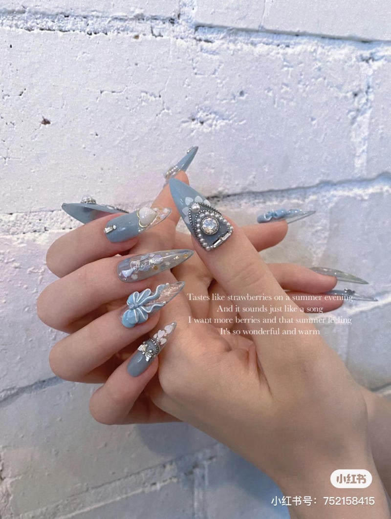 mau-nail-mong-nhon-dai__5__b018d5aa5bb94621aded9c338bc88069 Bàn tay với móng tay dài, trang trí màu xanh dương và họa tiết nổi bật, nền gạch trắng.