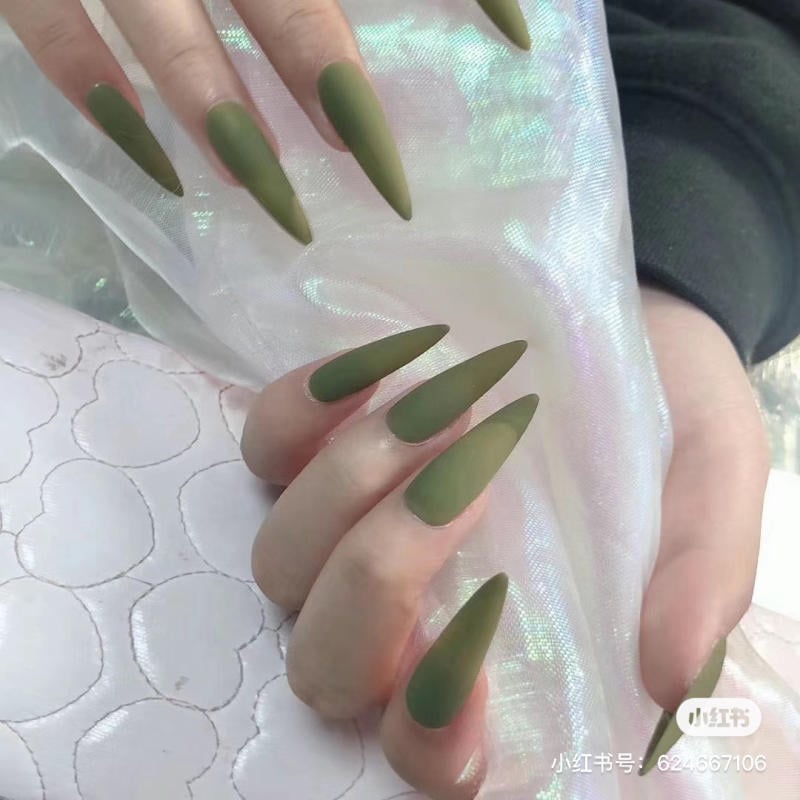 mau-nail-mong-nhon-dai__9__55701268552d4d3f9361fc39886cf7aa Bàn tay với móng tay dài, nhọn được sơn màu xanh lá cây mờ, đặt trên nền vải sáng bóng.