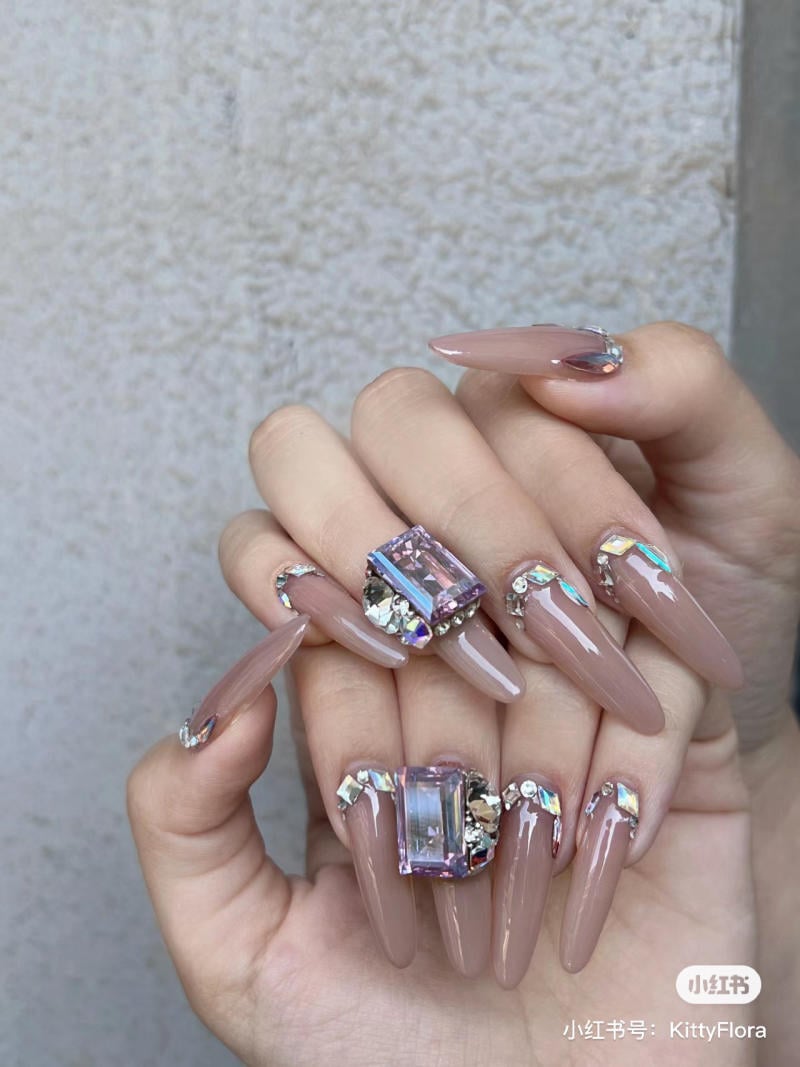 mau-nail-mong-nhon-tron__12__226756f0a61d409293f99b6fa5ee78a1 Bàn tay với móng dài, màu nâu, trang trí đá lấp lánh và họa tiết độc đáo trên nền tường sáng.