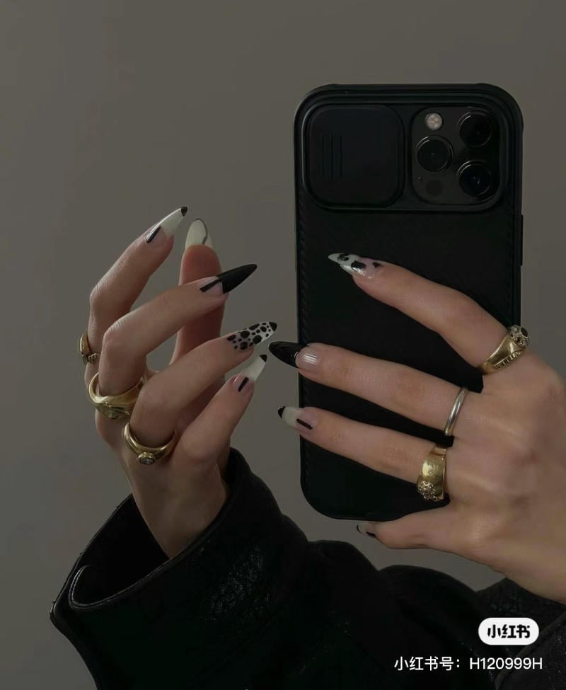 mau-nail-mong-nhon-tron__2__b46176dc4dc2430eb45c88a689a31726 Người đang cầm điện thoại đen, móng tay dài sơn họa tiết đen trắng, đeo nhiều nhẫn vàng và bạc.