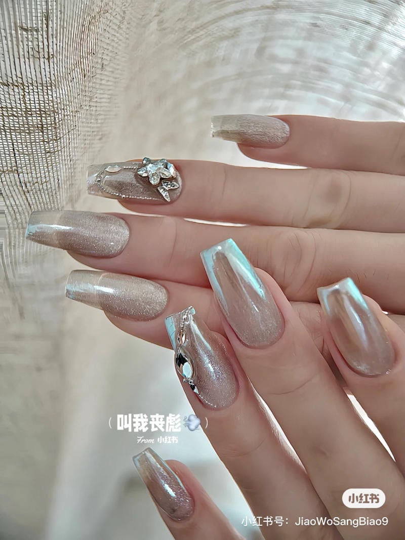 Bàn tay với móng tay dài sơn màu ánh kim, trang trí họa tiết và đá lấp lánh trên nền vải sáng.
