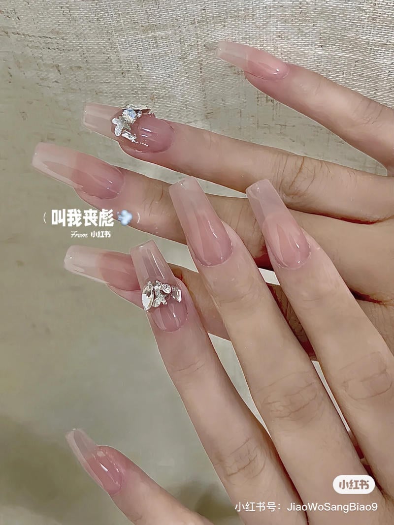 Bàn tay với móng tay dài màu hồng nhạt, đính đá lấp lánh trên một số móng.