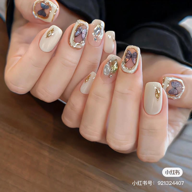 Bàn tay với móng tay được sơn màu nude, trang trí họa tiết hình bướm và đính đá lấp lánh trên bề mặt.