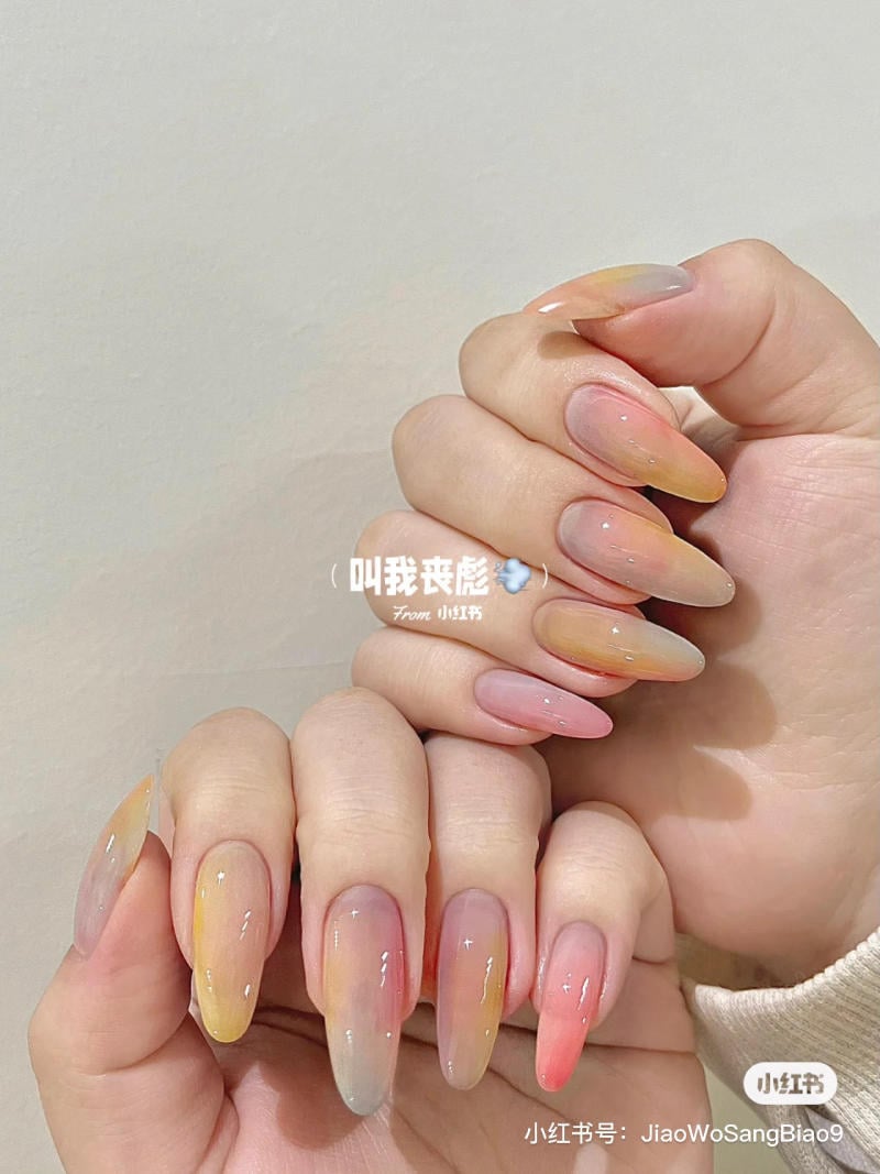 Hai bàn tay với móng tay dài sơn màu ombre hồng, vàng và xám, được chụp trên nền trắng.