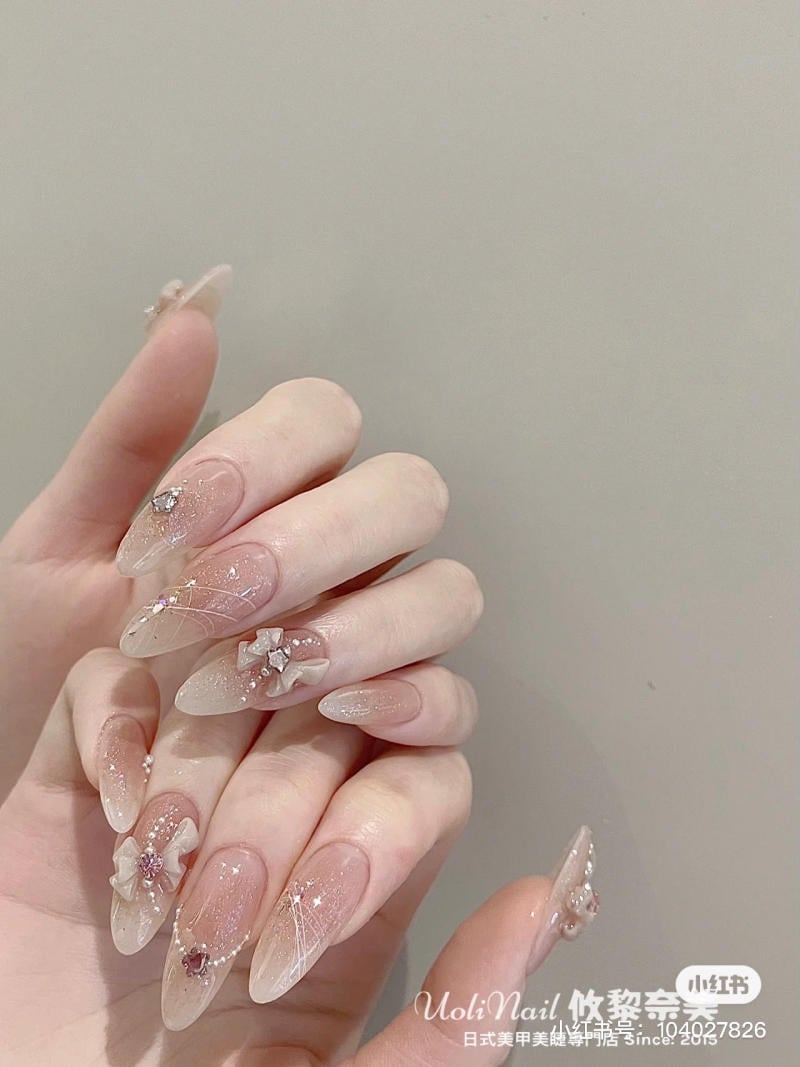 Bàn tay với móng tay dài, trang trí họa tiết hoa và kim tuyến lấp lánh, nền màu hồng nhạt.