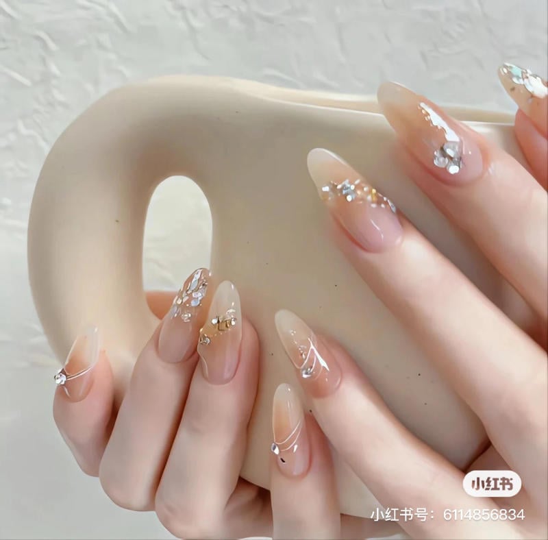 Bàn tay với móng tay dài, được trang trí bằng hoa văn và hạt đá lấp lánh, đang cầm một chiếc bình màu be.