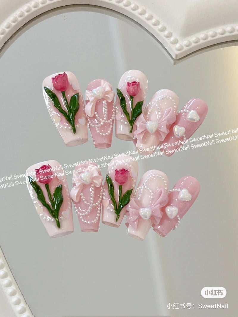Bộ móng giả màu hồng trang trí hoa hồng, nơ và ngọc trai trên nền gương phản chiếu.