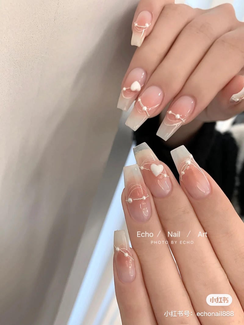 Bàn tay với móng tay dài, sơn màu hồng nhạt, trang trí hoa văn trắng và ngọc trai nhỏ.