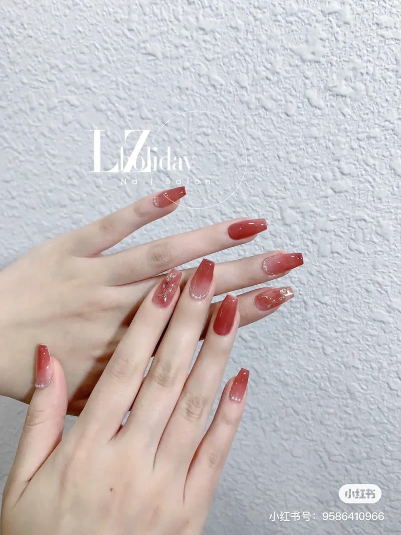 Bàn tay với móng tay màu đỏ bóng, có điểm nhấn trang trí vàng, đặt trên nền tường trắng.
