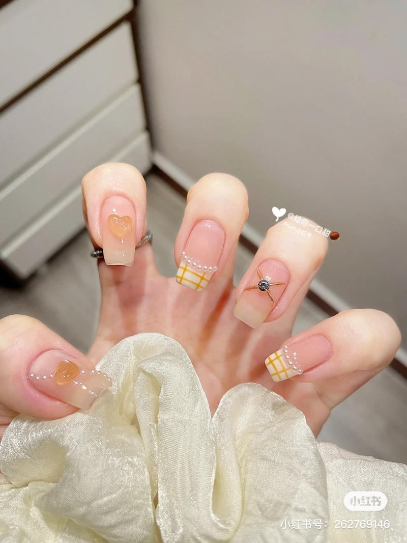 Bàn tay với móng tay được trang trí họa tiết trái tim, lưới kẻ và đính hạt, nền vải mềm mại màu trắng.